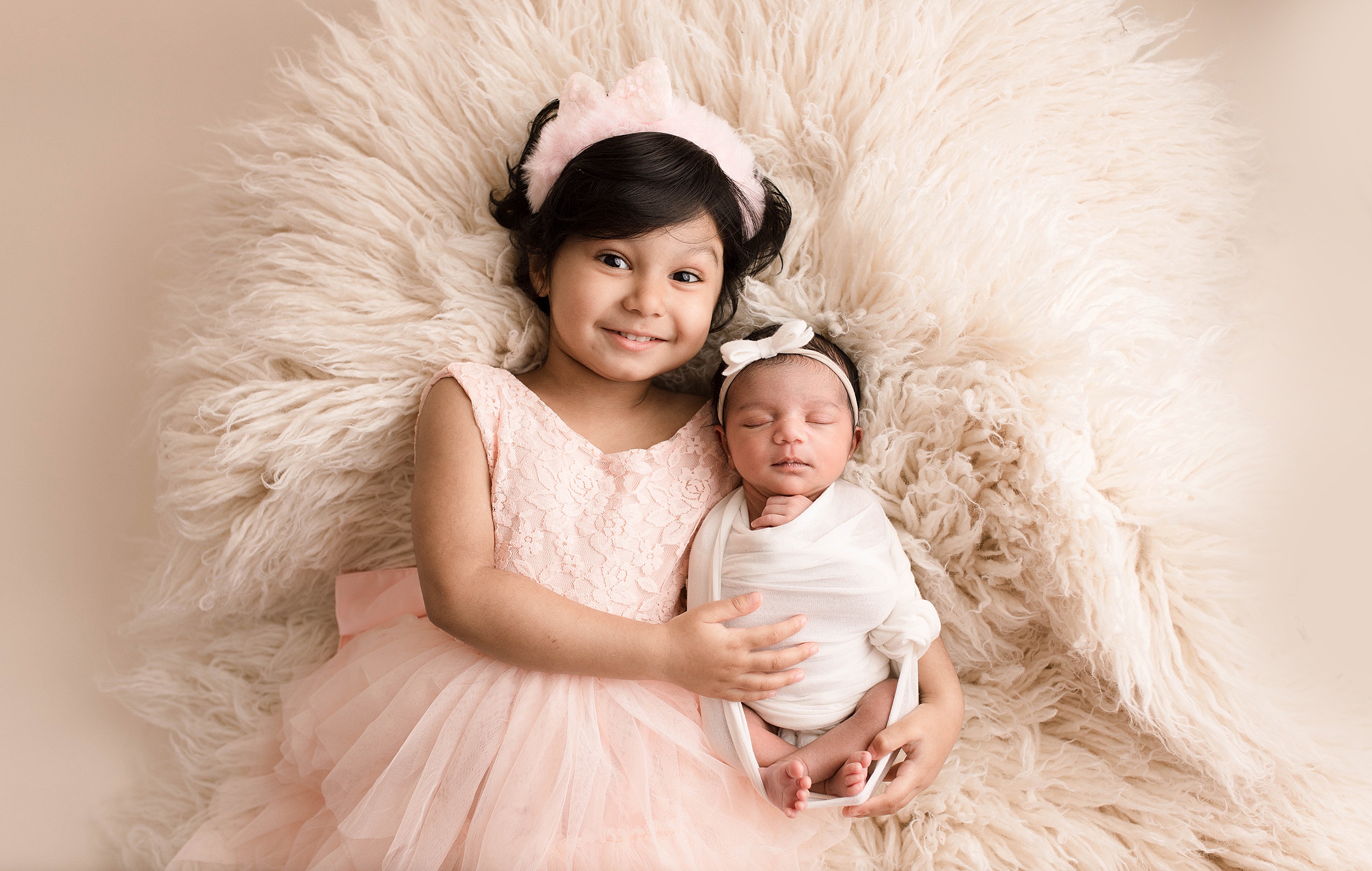 Young girl lays on a furry rug with newborn sister wearing a pink dress and headband Seattle-Pediatricians