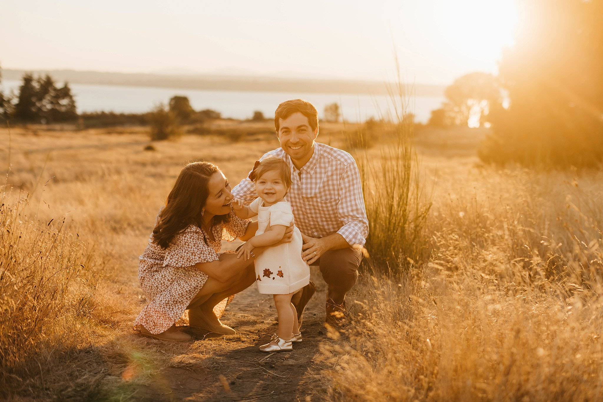 Mother and father play in a golden grassy field at sunset with their toddler daughter seattle prenatal massage